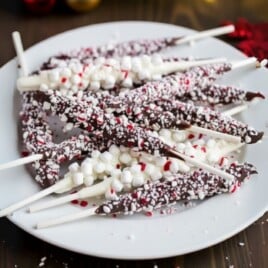White plate piled with chocolate and white hot chocolate stir sticks generously coated with crushed peppermint and mini marshmallows, styled on a wooden table with evergreen branches, red flowers, and glowing Christmas lights.