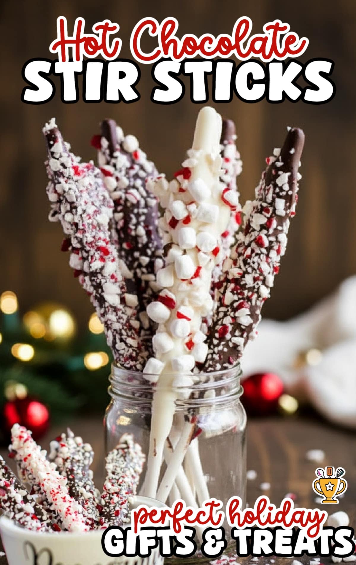 Clear mason jar filled with festive peppermint hot chocolate stir sticks coated in chocolate and white almond bark, covered with crushed peppermint and mini marshmallows, styled with Christmas ornaments and blurred holiday background.