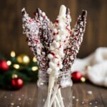 Glass mason jar filled with peppermint hot chocolate stir sticks coated in chocolate and white almond bark, topped with crushed peppermint and mini marshmallows, styled with red and gold ornaments and soft holiday lights in the background.