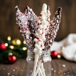 Glass mason jar filled with peppermint hot chocolate stir sticks coated in chocolate and white almond bark, topped with crushed peppermint and mini marshmallows, styled with red and gold ornaments and soft holiday lights in the background.