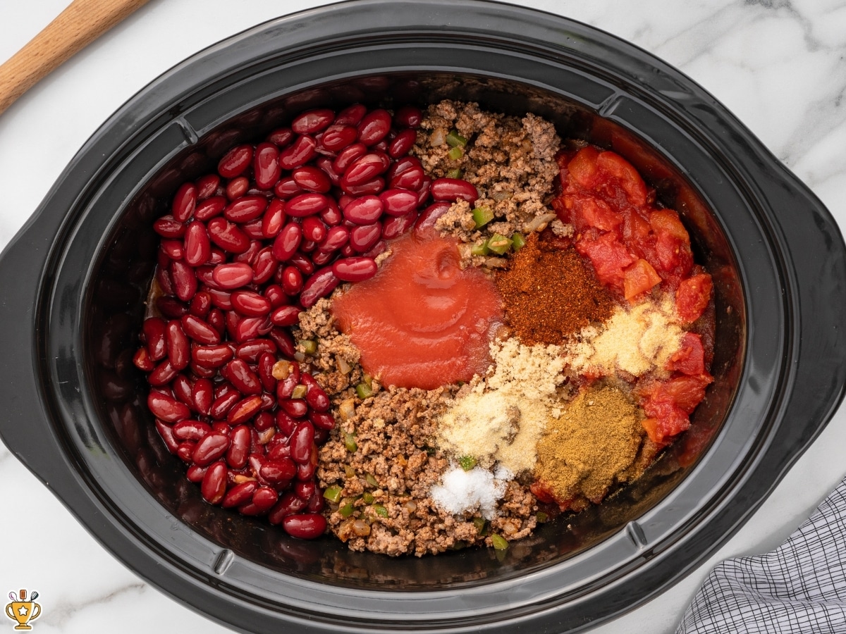 Browned beef, kidney beans, diced tomatoes, tomato sauce, and seasonings added to a crockpot before cooking chili.
