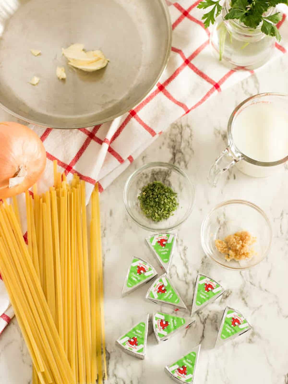 Ingredients for skinny Laughing Cow alfredo sauce laid out on a marble surface, including pasta, Laughing Cow cheese wedges, milk, chives, garlic, and onion.