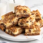 Stack of chocolate chip cheesecake bars on a white plate, showing thick cookie dough layers, creamy cheesecake filling, and melty chocolate chips with glasses of milk in the background.