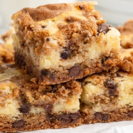 Chocolate chip cheesecake bars stacked on a white plate, highlighting the golden cookie dough top, creamy cheesecake center, and chocolate-studded cookie crust.