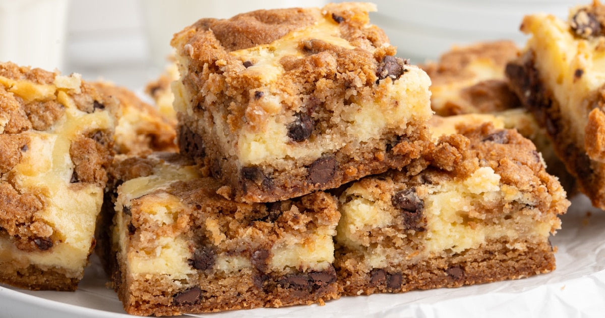 Chocolate chip cheesecake bars stacked on a white plate, highlighting the golden cookie dough top, creamy cheesecake center, and chocolate-studded cookie crust.