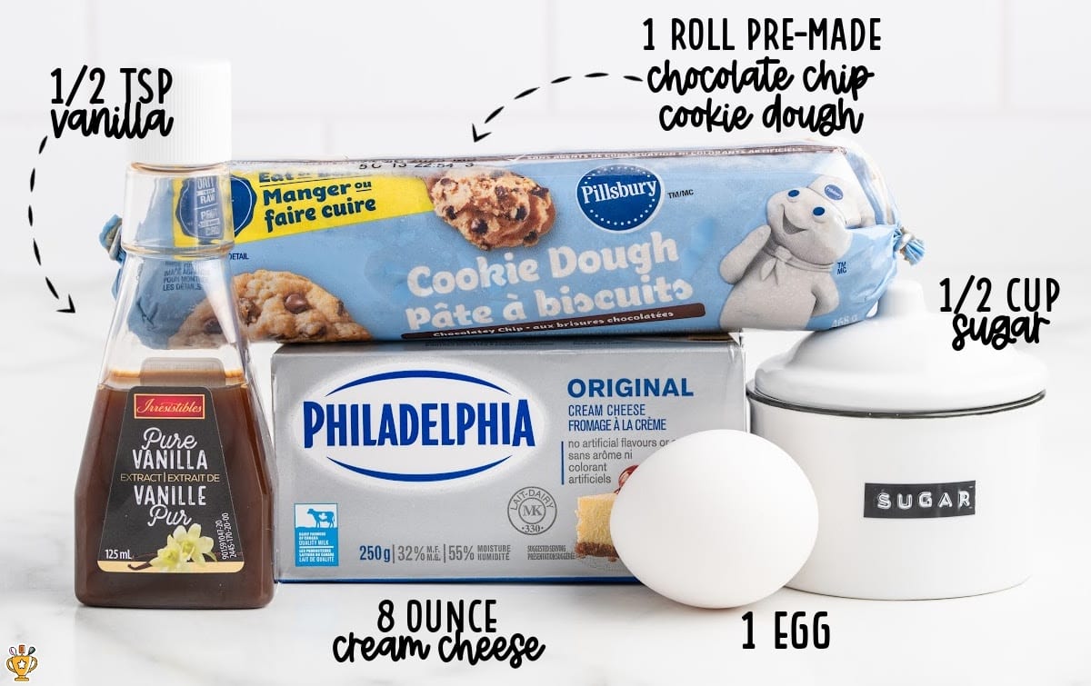 Labeled ingredients for chocolate chip cheesecake bars, including cookie dough, cream cheese, sugar, vanilla, and an egg displayed on a white surface.