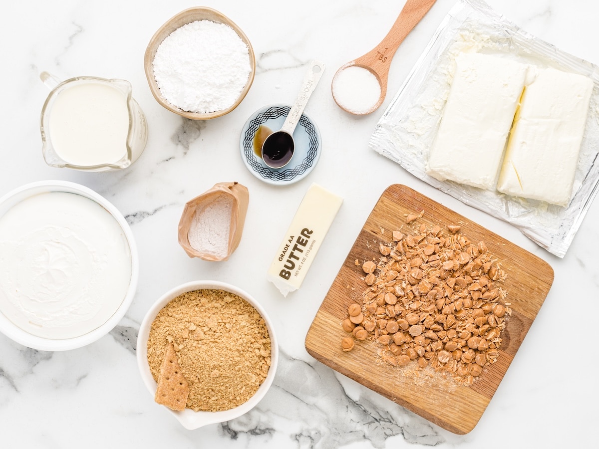 Overhead view of Butterscotch Delight ingredients on marble: cream, powdered sugar, cream cheese, vanilla, whipped topping, pudding mix, butter, graham cracker crumbs, and chopped butterscotch chips.