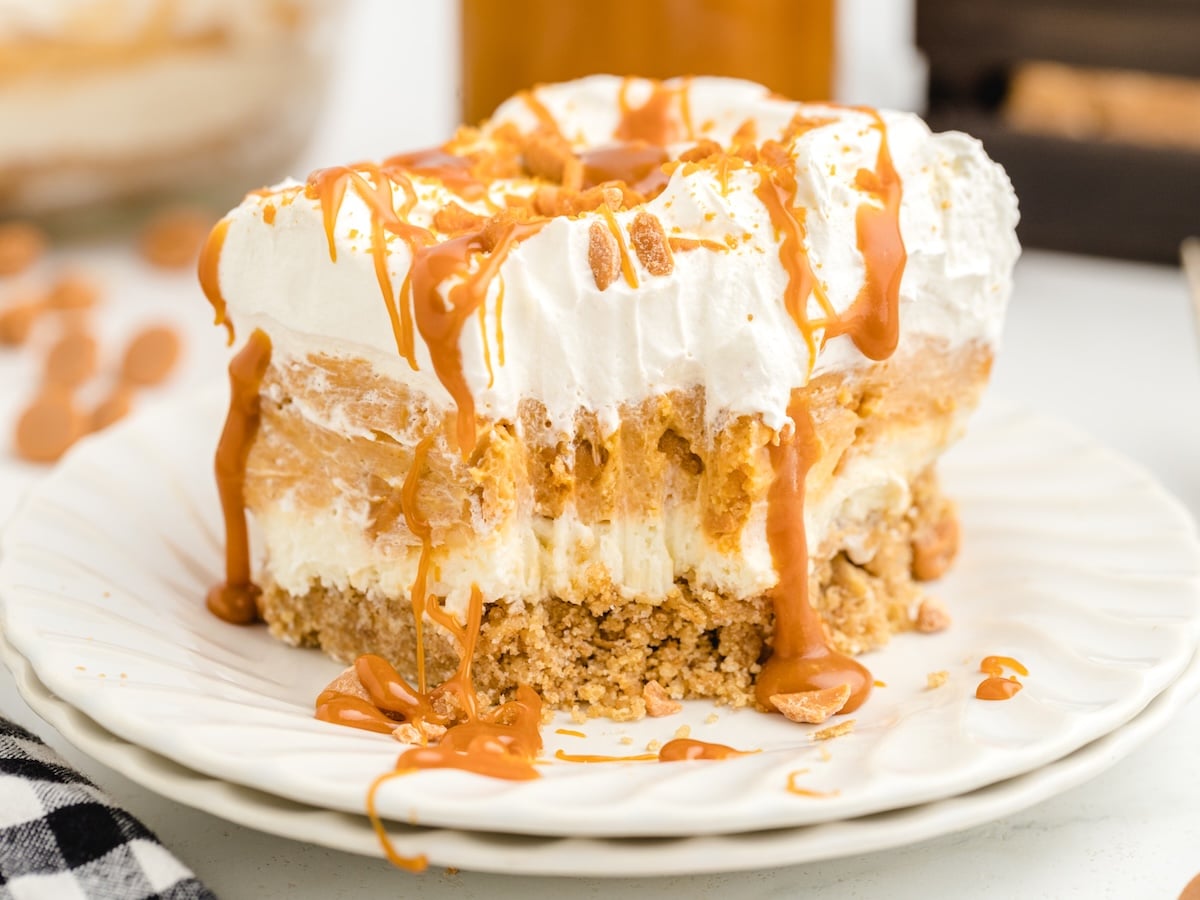 Square slice of no-bake Butterscotch Delight on a white plate, showing graham cracker crust, cream cheese layer, butterscotch pudding, and whipped topping, with butterscotch sauce dripping down the sides.
