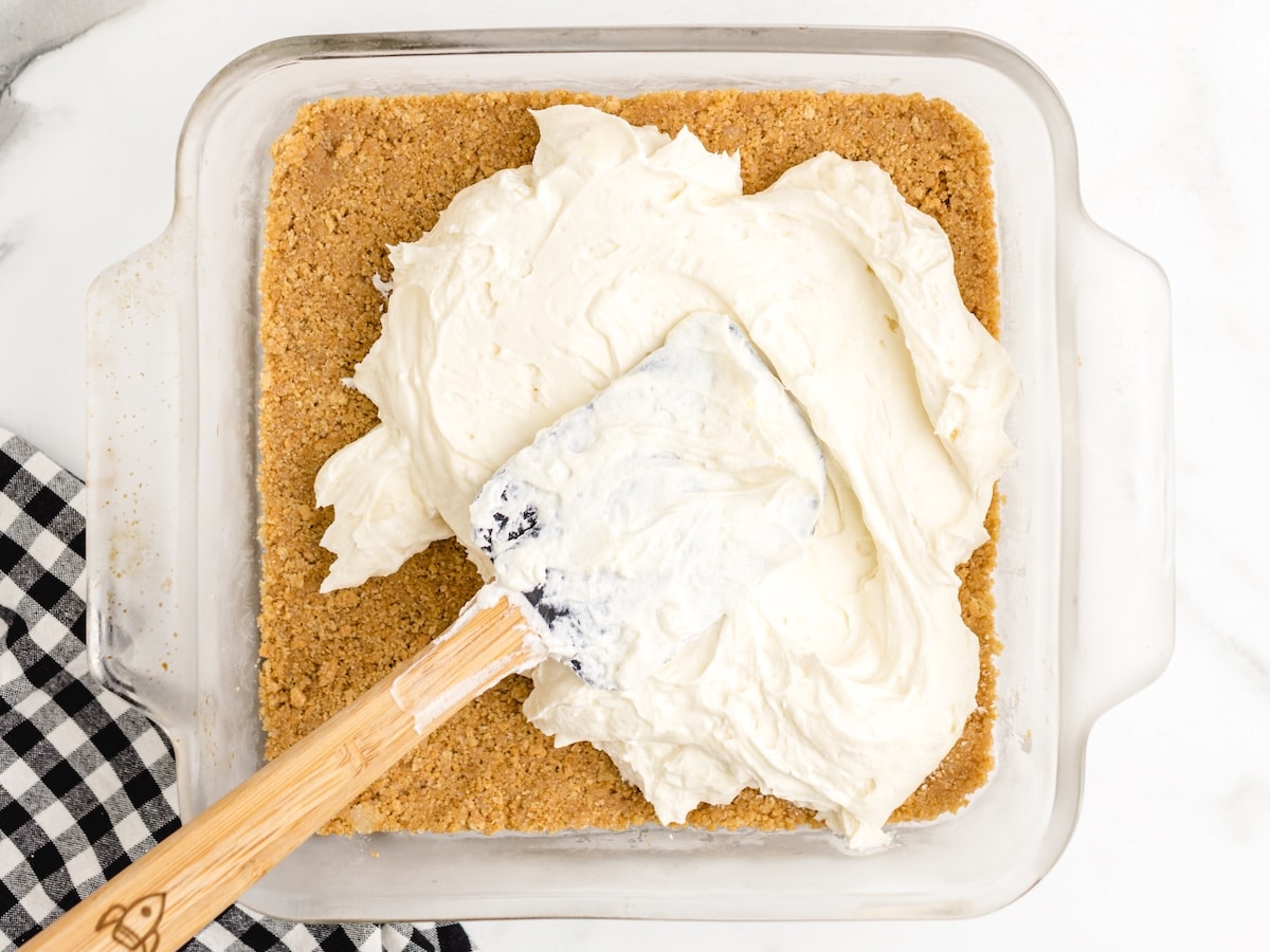 Spreading half of the cream cheese layer over the graham cracker crust with a spatula.