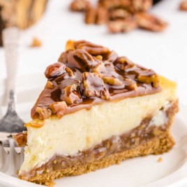 A plate contains a piece of Pecan Pie Cheesecake with a fork.