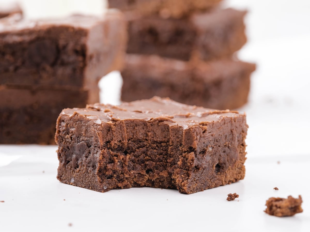 Rich, fudgy lunch lady brownies with a glossy chocolate frosting layer, stacked brownie squares in the background, and a close-up brownie showing the dense, chewy texture inside.