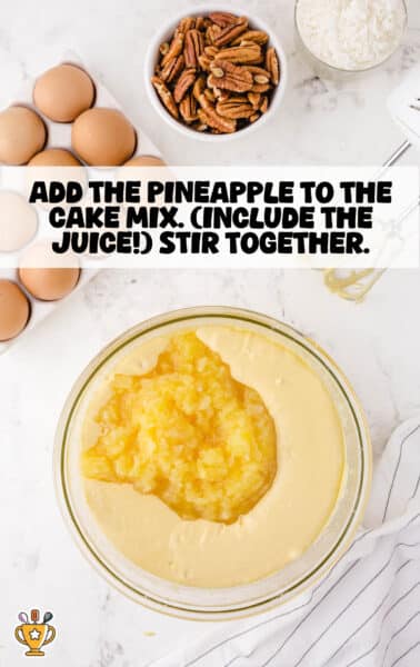 Overhead shot of a mixing bowl filled with yellow cake batter and crushed pineapple, surrounded by baking ingredients like eggs, pecans, and coconut on a white marble countertop.