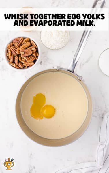 Metal saucepan filled with evaporated milk and two raw egg yolks, ready to be whisked together, surrounded by bowls of chopped pecans, shredded coconut, and sugar on a white marble countertop.