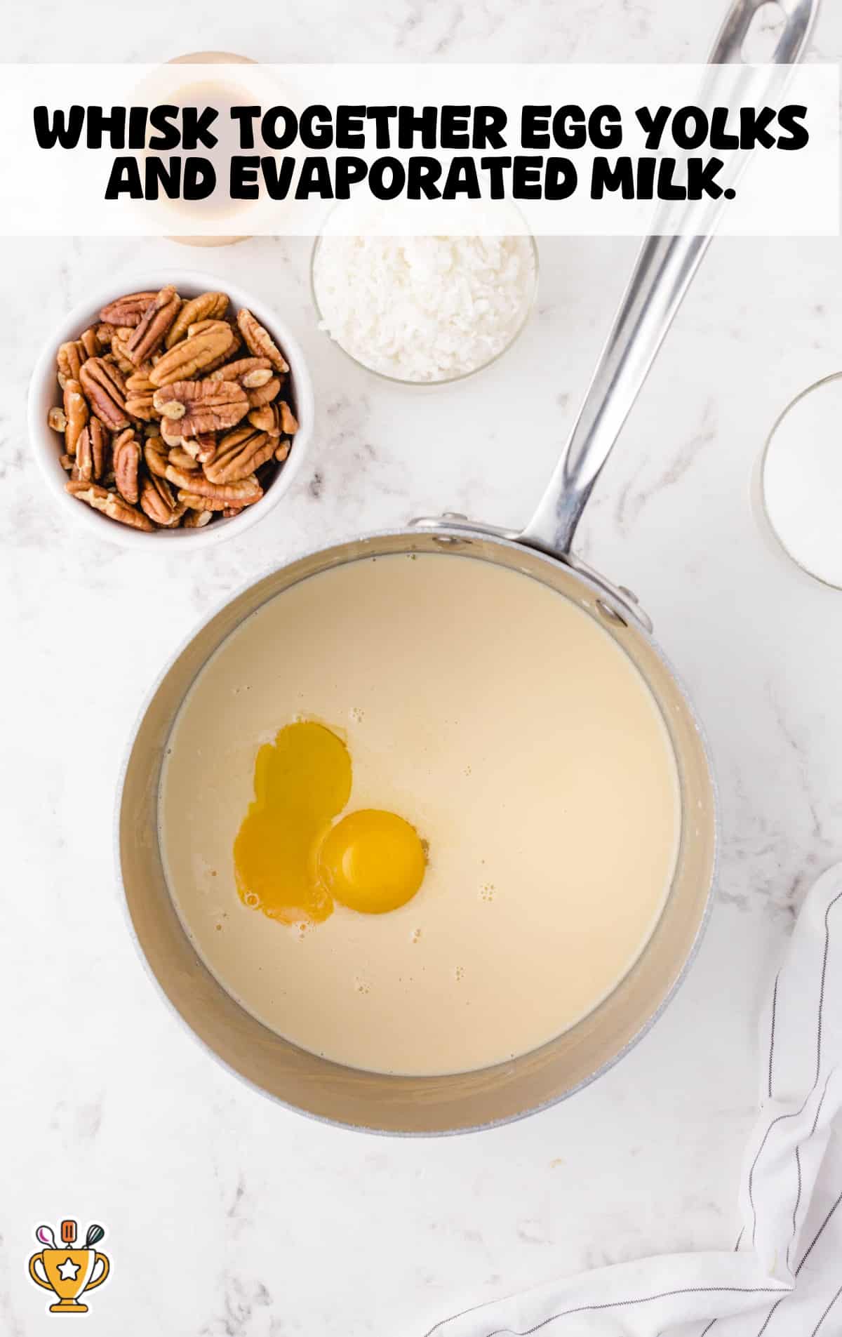 Metal saucepan filled with evaporated milk and two raw egg yolks, ready to be whisked together, surrounded by bowls of chopped pecans, shredded coconut, and sugar on a white marble countertop.