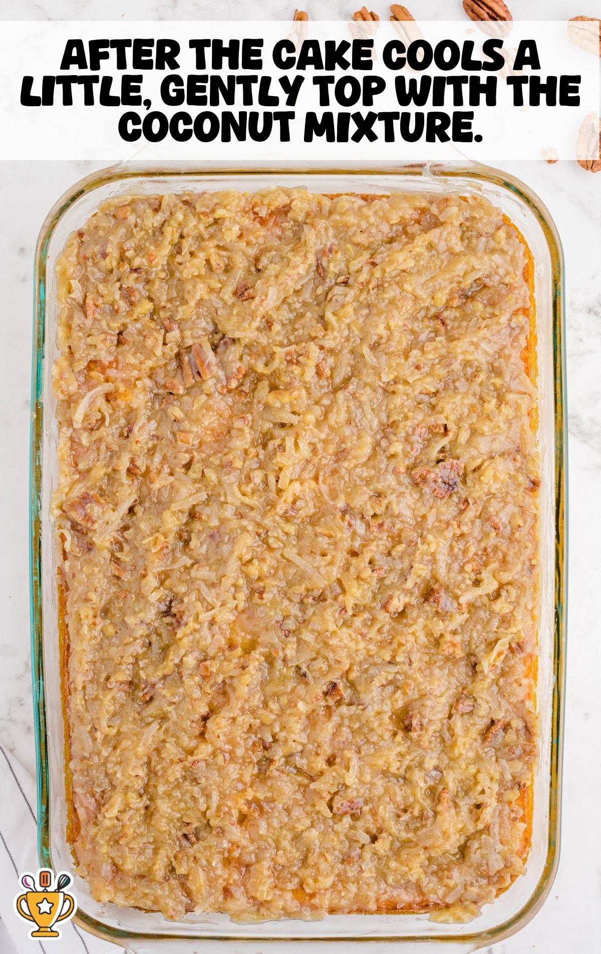 Top-down view of a glass baking dish filled with baked Southern-style tornado cake topped with a thick layer of golden brown coconut and pecan mixture, resting on a white marble surface with scattered pecans in the background.