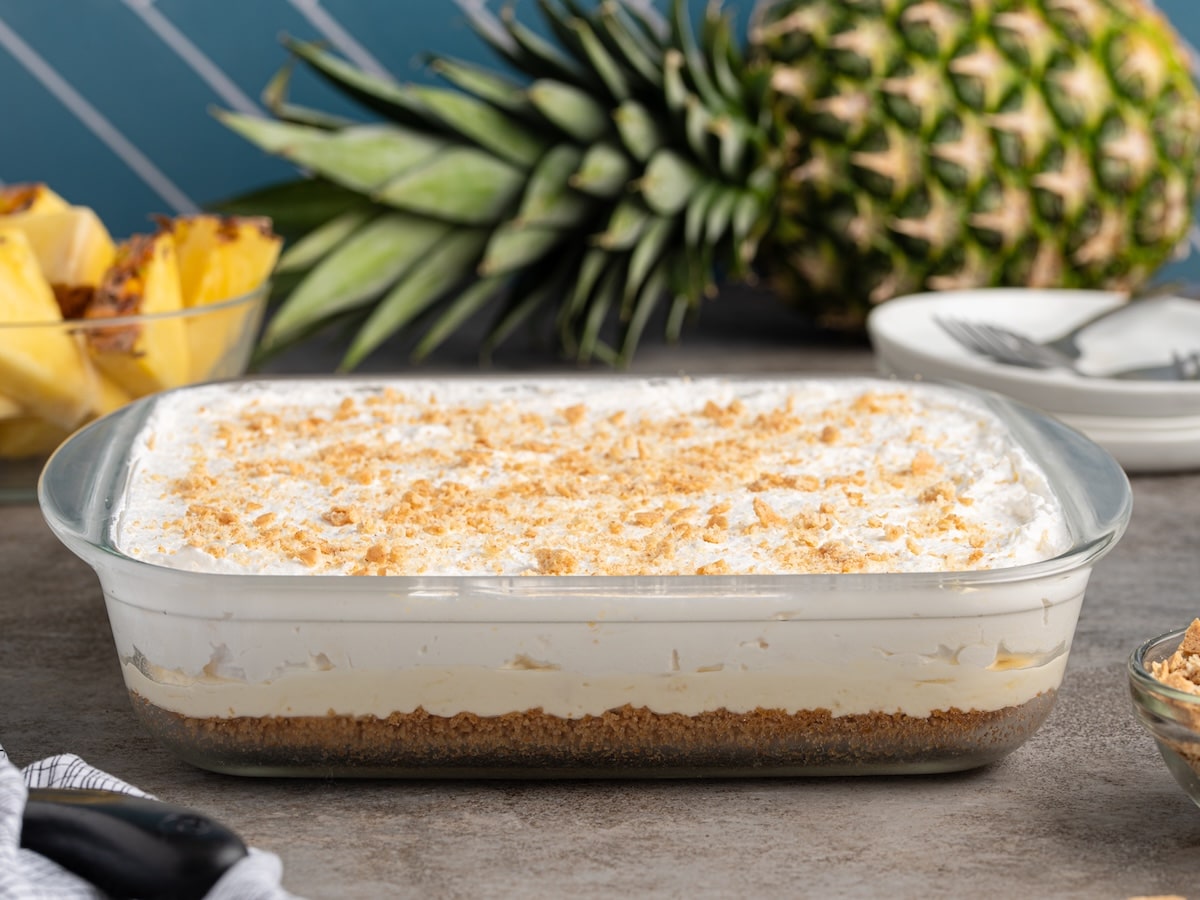 Pineapple delight layered dessert with graham cracker crust, creamy pineapple filling, and whipped topping sprinkled with graham cracker crumbs.