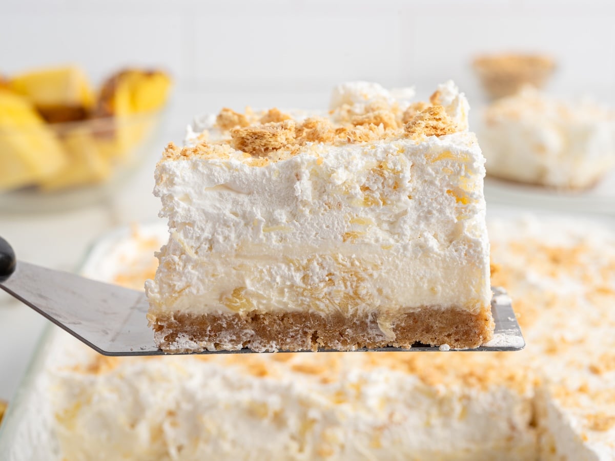 Slice of pineapple delight on a spatula showing layers of graham cracker crust, creamy pineapple filling, and whipped topping.