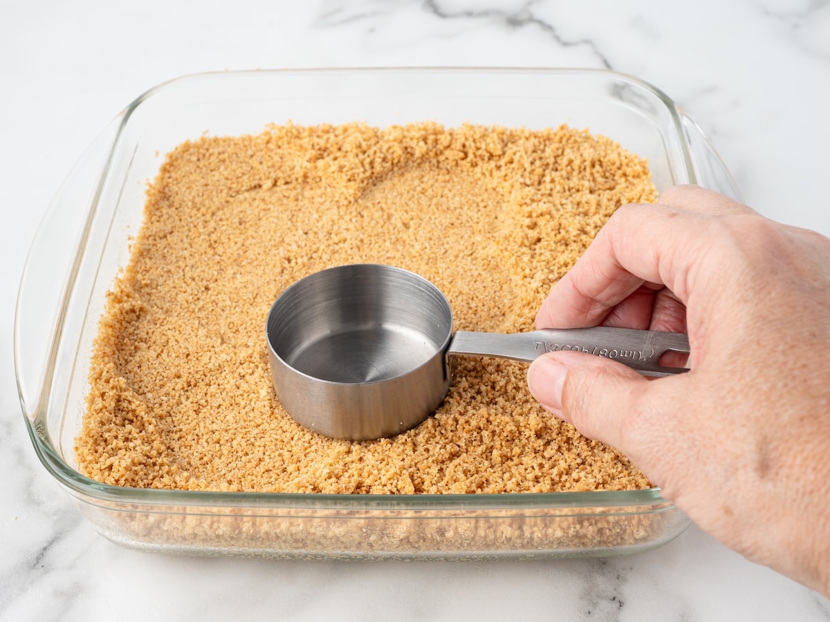 Graham cracker crust being pressed into a glass baking dish with a measuring cup.