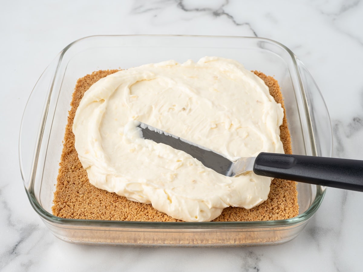 Cream cheese filling being spread over a baked graham cracker crust in a glass baking dish.