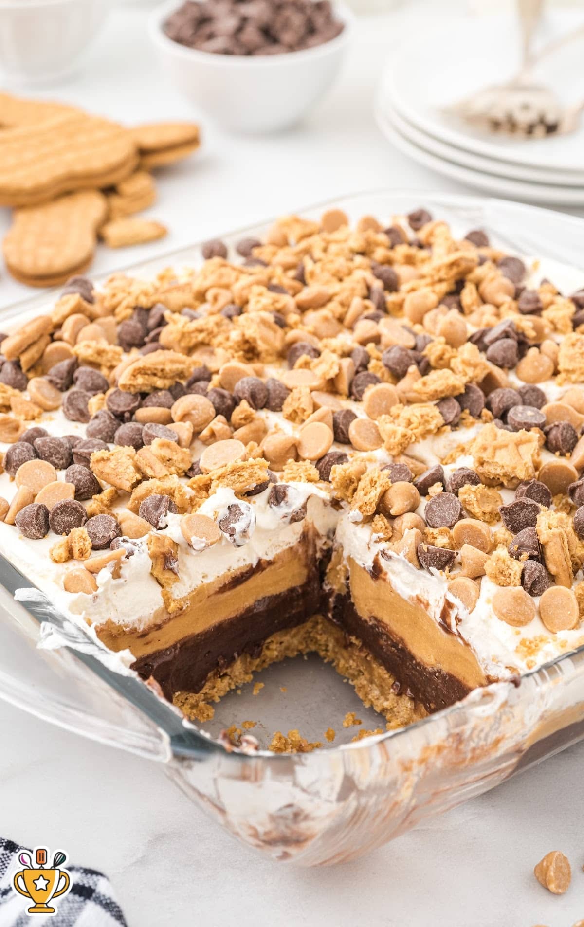 Glass baking dish filled with peanut butter dream bars showing neat dessert layers&mdash;cookie crust, chocolate pudding, peanut butter layer, and whipped topping&mdash;topped with crushed cookies and chocolate chips. A no-bake peanut butter chocolate dessert.