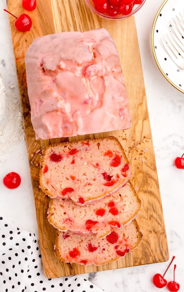 A piece of cake sitting on top of a wooden cutting board, with Cherry and Bread