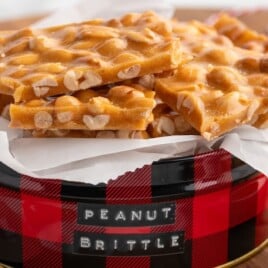 Stack of golden peanut brittle pieces arranged in a red buffalo-plaid holiday tin lined with parchment paper, highlighting the rich caramel color and whole roasted peanuts.