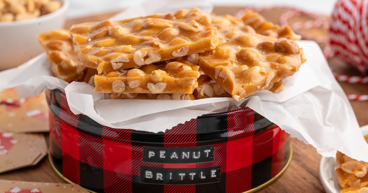 Stack of golden peanut brittle pieces arranged in a red buffalo-plaid holiday tin lined with parchment paper, highlighting the rich caramel color and whole roasted peanuts.