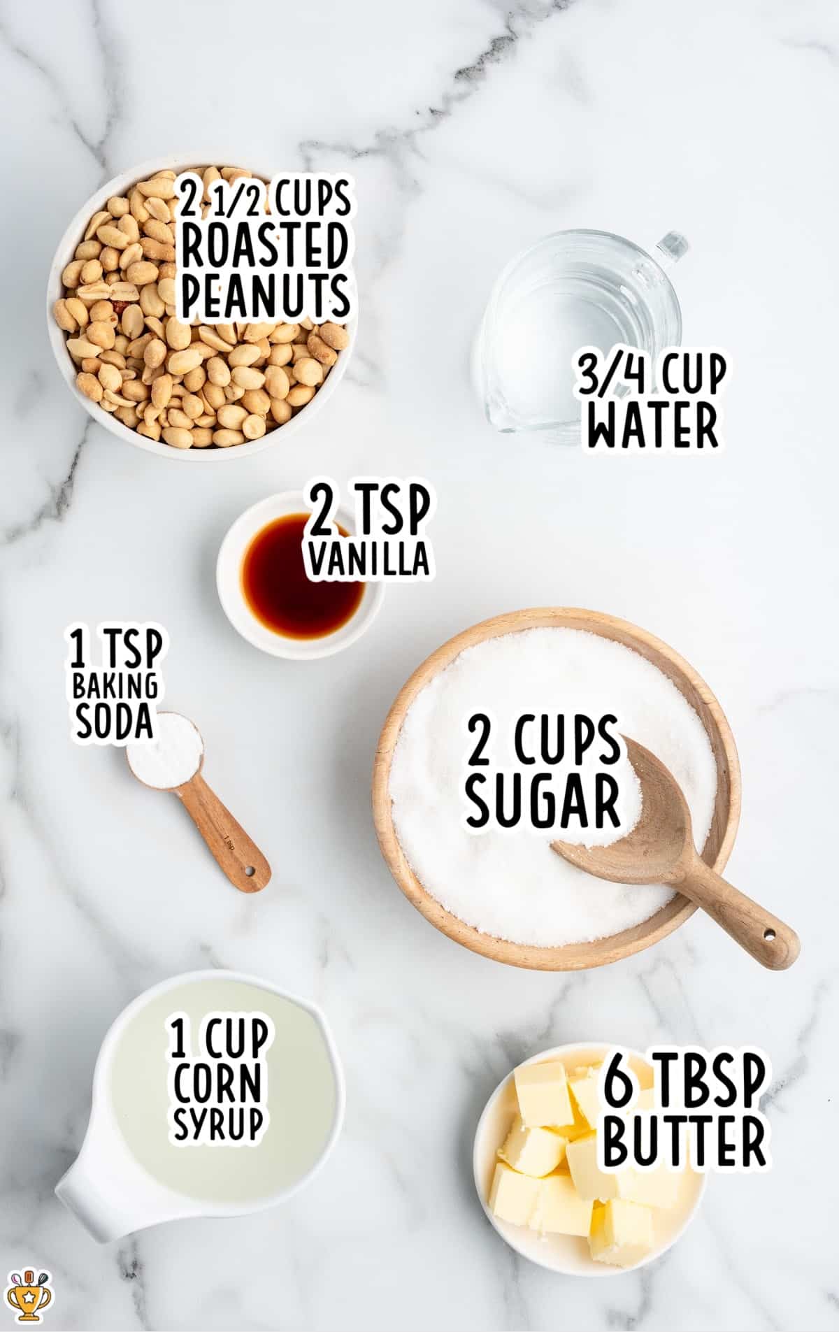Labeled peanut brittle ingredients on a marble surface: roasted peanuts, sugar, corn syrup, butter, vanilla, baking soda, and water displayed in bowls and measuring cups.