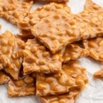 Pile of homemade peanut brittle squares on white parchment paper, showcasing the smooth caramel snap and roasted peanuts throughout each piece.