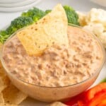 Crockpot Rotel dip being scooped with a tortilla chip, displayed above a clear ingredient list featuring Velveeta, Rotel tomatoes, and ground beef.