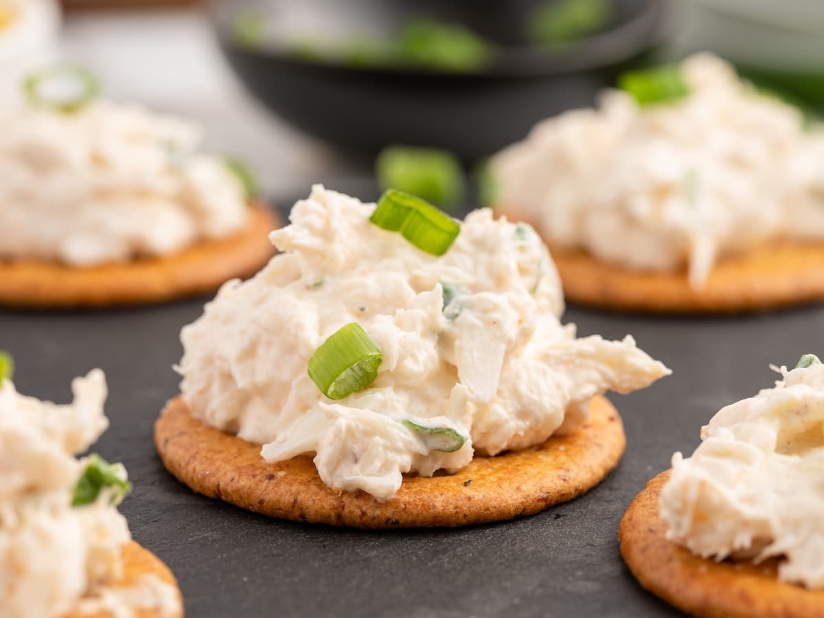 Creamy cold crab dip topped with sliced green onions served on round crackers arranged on a dark serving board. The dip has visible pieces of lump crab meat and a smooth cream cheese base.