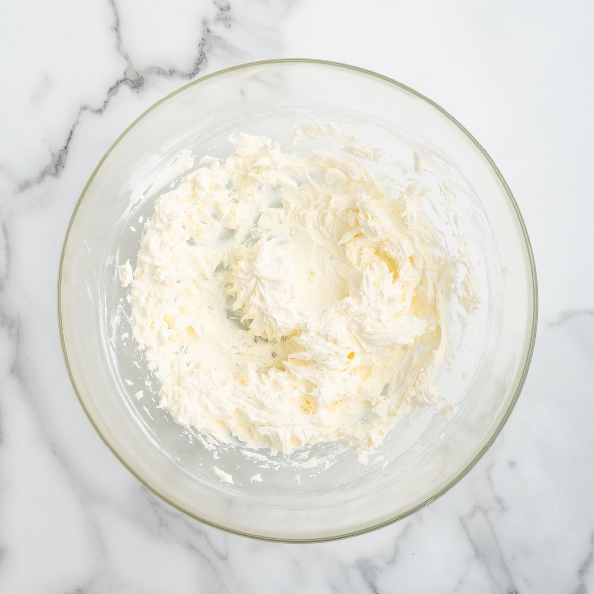 Softened cream cheese whipped until smooth and fluffy in a clear glass mixing bowl on a white marble surface, ready to be combined with the remaining cold crab dip ingredients.