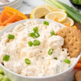 Creamy cold crab dip garnished with sliced green onions in a white serving bowl, surrounded by crackers, celery sticks, bell pepper slices, lemon wedges, and fresh green onions.