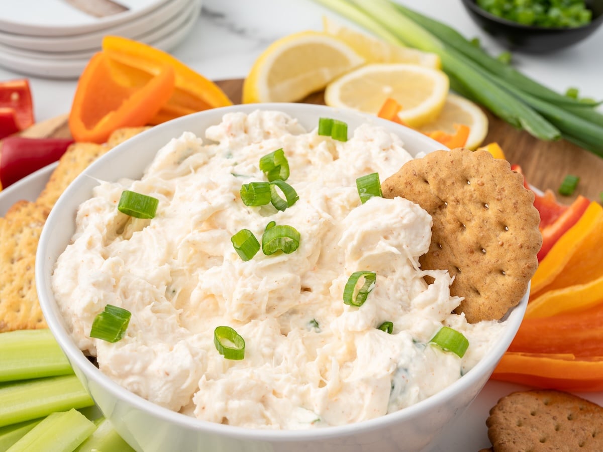Creamy cold crab dip garnished with sliced green onions in a white serving bowl, surrounded by crackers, celery sticks, bell pepper slices, lemon wedges, and fresh green onions.