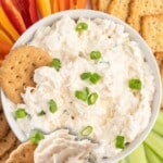 A cracker topped with creamy cold crab dip is held in front of a white bowl filled with crab dip and garnished with sliced green onions. Crackers, celery sticks, and colorful bell pepper strips surround the bowl.
