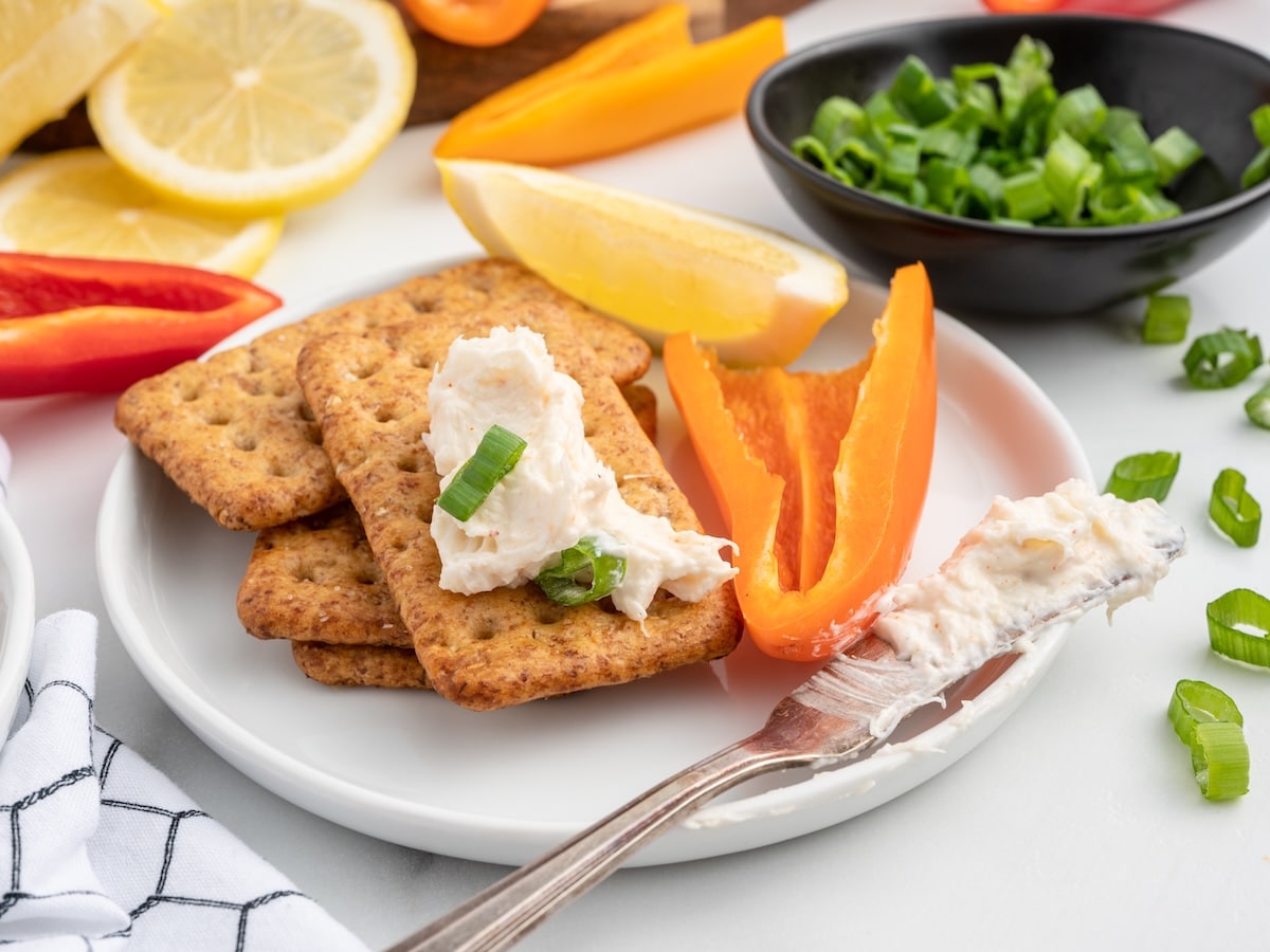 Crackers topped with cold crab dip sit on a white plate alongside sliced bell peppers and lemon wedges. A butter knife spread with crab dip rests on the plate, with sliced green onions scattered around the serving area.