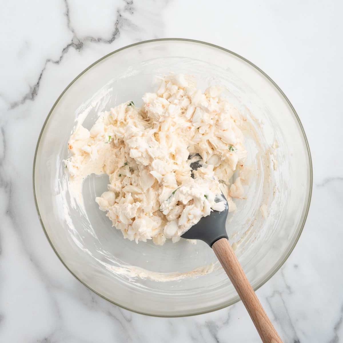Lump crab meat gently folded into a seasoned cream cheese mixture in a glass mixing bowl with a silicone spatula, creating a chunky and creamy cold crab dip mixture.