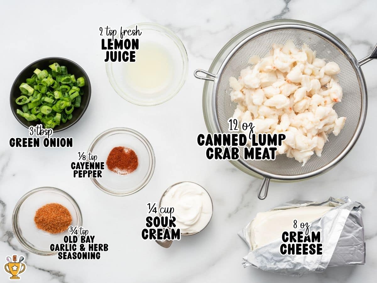 Ingredients for cold crab dip arranged on a white marble surface, including cream cheese, sour cream, drained lump crab meat, sliced green onions, fresh lemon juice, Old Bay Garlic & Herb Seasoning, and cayenne pepper measured in small bowls