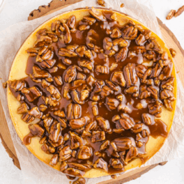 Overhead view of a full pecan pie cheesecake on parchment paper and a wooden board, topped with caramel sauce and a generous layer of glossy pecans — perfect holiday dessert combining creamy cheesecake and classic pecan pie flavors.
