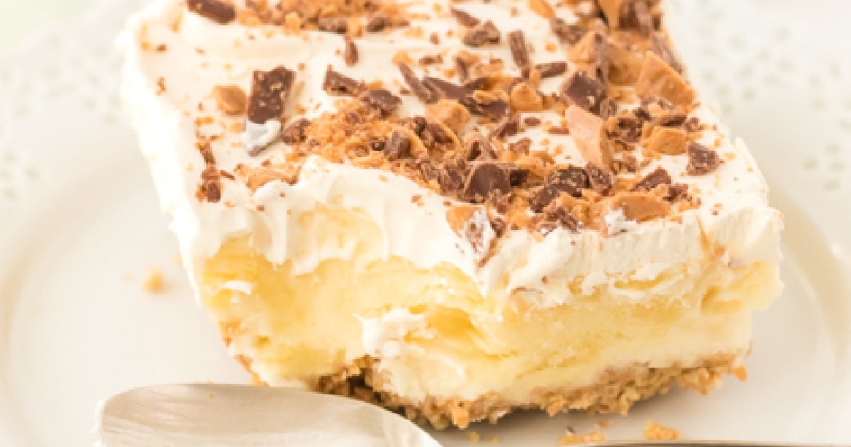 Close-up slice of no-bake butter pecan crunch dessert showing creamy vanilla pudding layers, whipped topping, and crushed Heath bar pieces on a graham cracker crust.