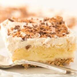 Close-up of a thick, creamy slice of butter pecan crunch no-bake dessert on a plate, showing the graham cracker crust, pudding layer, and whipped topping with crunchy toffee pecans.