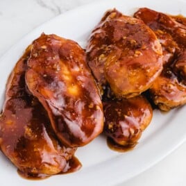 Close-up of four juicy Coca Cola chicken breasts coated in thick, glossy sauce, served on a white oval platter.