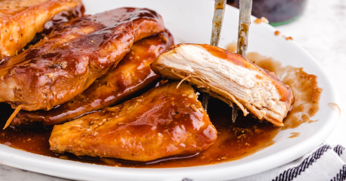 Close-up of Coca Cola chicken on a white serving plate, showcasing moist, saucy chicken breasts with a fork holding a piece to show the tender texture.