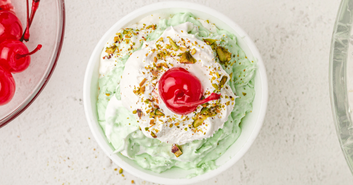 close up shot of a small bowl of pistachio salad topped with whipped cream and crushed pistachios with a cherry on top