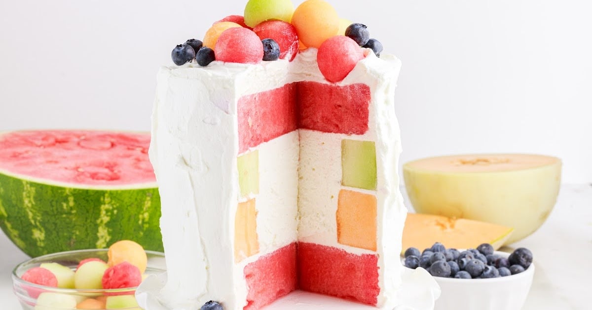 Watermelon Cake on a cake stand with a slice taken out and put on a plate topped with melon balls and blueberries