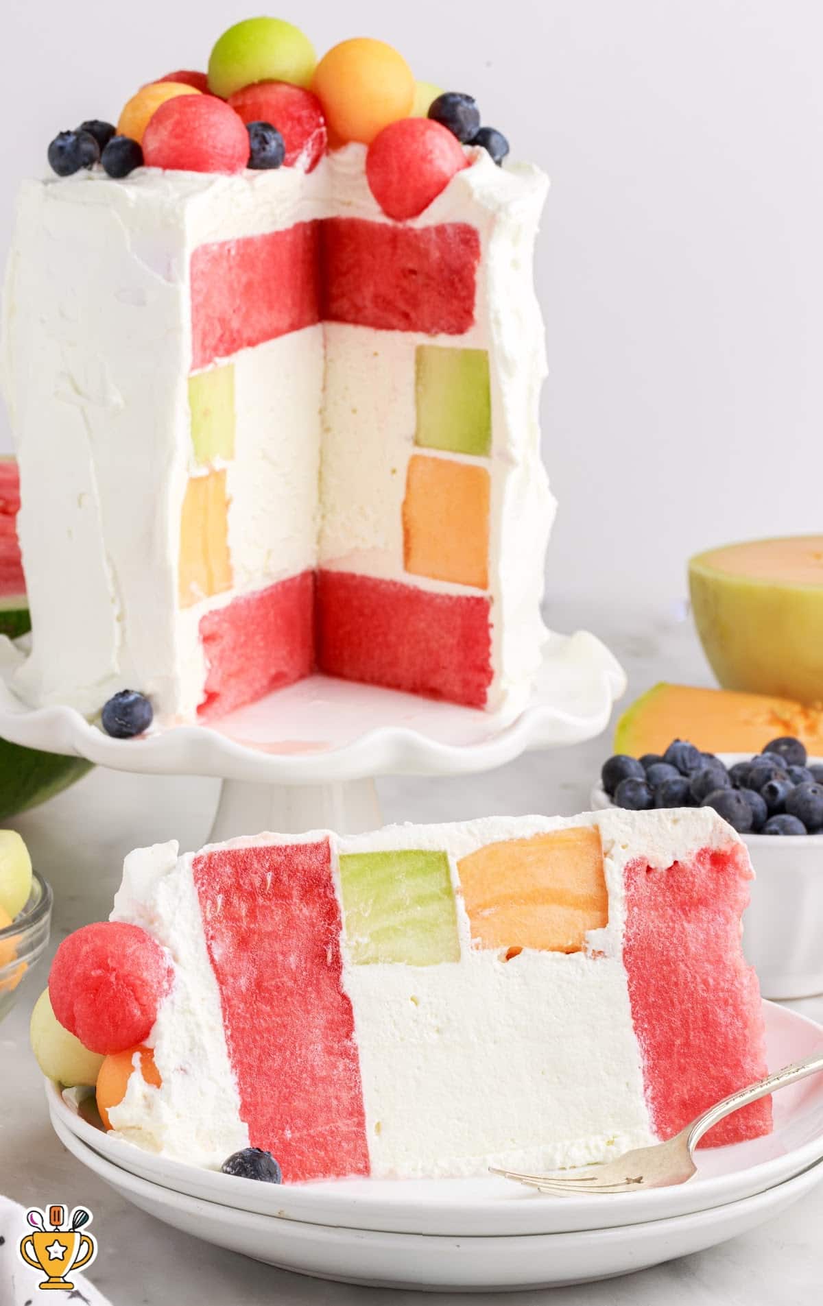 Watermelon Cake on a cake stand with a slice taken out and put on a plate topped with melon balls and blueberries