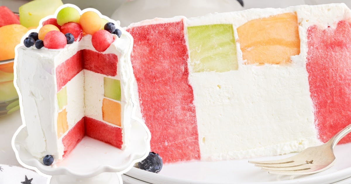 Watermelon Cake on a cake stand with a slice taken out and put on a plate topped with melon balls and blueberries