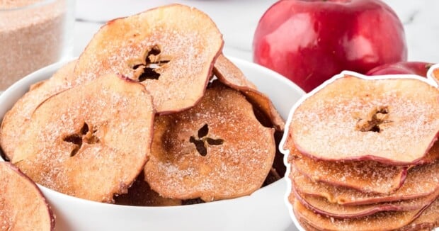 a bowl of apple chips next to a stack of apples with cinamon 