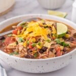 a bowl of chicken taco soup garnished with cheese and lime wedges.