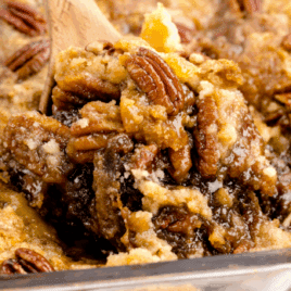 Close-up of a warm pecan dump cake being scooped from a glass baking dish, showing gooey brown sugar filling, golden cake mix topping, and whole toasted pecans.